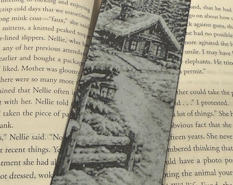 May include: Black and white bookmark featuring a detailed illustration of a snow-covered cabin nestled among pine trees and mountains. The bookmark is rectangular and is placed between the pages of a book.