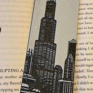 May include: A rectangular bookmark featuring a detailed black and white illustration of a city skyline, including a prominent skyscraper. The bookmark is made of a light gray material and is placed on top of an open book.