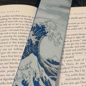 May include: A bookmark featuring a blue and white wave design, reminiscent of the iconic Great Wave off Kanagawa. The bookmark is rectangular and is placed in a book with visible text.
