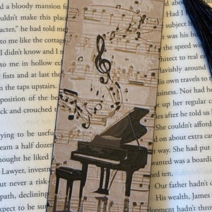 May include: A bookmark with a musical theme. The bookmark features a black grand piano and musical notes on a beige background. A black tassel hangs from the top. The background has musical notes printed on it.