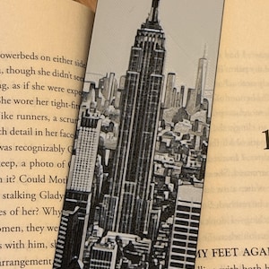 May include: A black and white bookmark featuring a detailed illustration of the New York City skyline, prominently showcasing the Empire State Building. The bookmark is rectangular and is placed inside an open book.