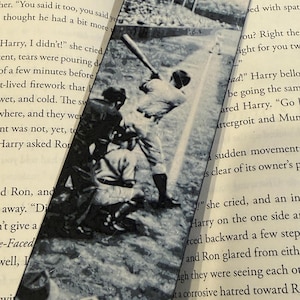 May include: Black and white baseball-themed bookmark featuring a vintage photograph of a baseball game. The image shows a batter swinging at a pitch. The bookmark is rectangular and is placed on top of an open book.