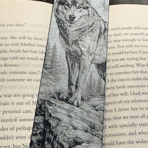 Puede incluir: Un marcap&aacute;ginas en blanco y negro con una ilustraci&oacute;n detallada de un lobo sobre una superficie rocosa, con una monta&ntilde;a de fondo. El marcap&aacute;ginas est&aacute; insertado en un libro, con texto visible en ambos lados.