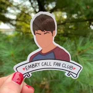 May include: A sticker featuring a cartoon illustration of a person with brown hair and a blue and maroon shirt. The sticker has a banner with the text "EMBRY CALL FAN CLUB" and two small red hearts.