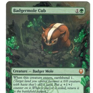 Könnte beinhalten: Eine Magic: The Gathering-Karte mit dem Titel "Badgermole Cub". Die Karte zeigt ein braun-weißes, dachsbartiges Wesen in einer Waldszene. Der Text beschreibt die Fähigkeiten und Werte der Kreatur, mit Stärke/Widerstand von 2/2.