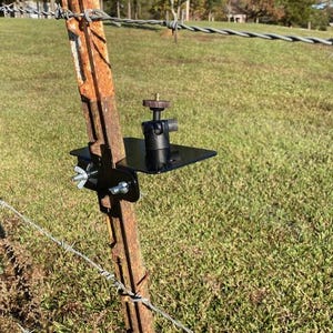 May include: A black metal camera mount attached to a rusty metal fence post. The mount has a small black platform and a swiveling ball head.
