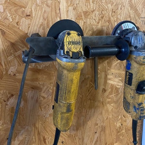 May include: Two yellow and black DeWalt angle grinders are mounted on a metal rack. The grinders have black guards and are attached to a wooden wall.