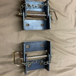 May include: Two metal winch brackets with a dark gray finish. Each bracket has a chain, a pin, and a gold-colored handle. The brackets are designed to be mounted and used for securing items.