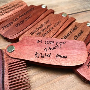 May include: A set of wooden pocket combs with personalized engravings. The combs are made of reddish-brown wood and have a brass hinge. The engravings include messages like "Happy 5th Anniversary", "Best Daddy Ever", and "We love you daddy!"