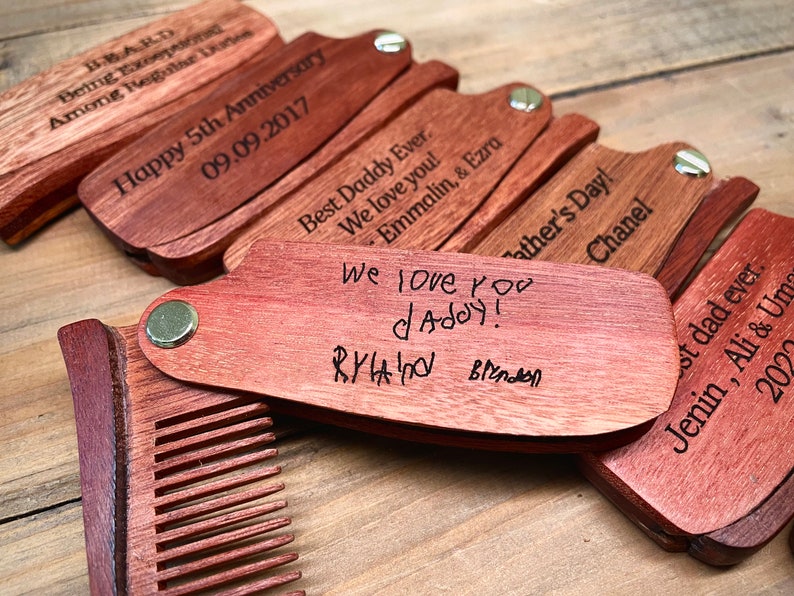 May include: A set of wooden pocket combs with personalized engravings. The combs are made of reddish-brown wood and have a brass hinge. The engravings include messages like "Happy 5th Anniversary", "Best Daddy Ever", and "We love you daddy!"