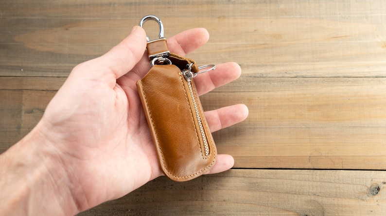 Pu&ograve; includere: Un portachiavi in ​​pelle marrone con chiusura a zip e un gancio in metallo argentato. Il portachiavi &egrave; tenuto in una mano.
