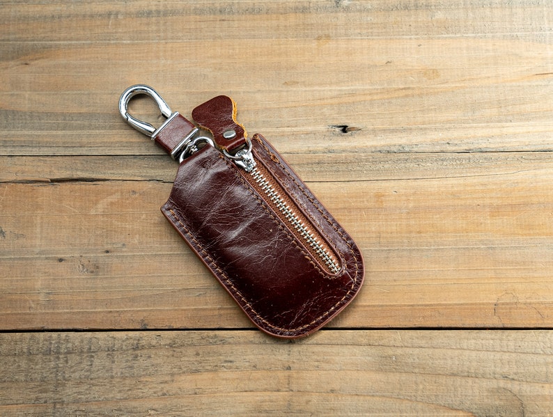Pu&ograve; includere: Un portachiavi in ​​cuoio marrone con una tasca con cerniera e un gancio in metallo argentato.