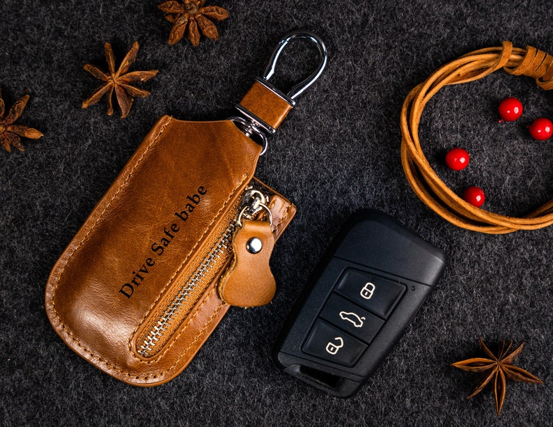 Pu&ograve; includere: Un portachiavi in ​​cuoio marrone con una tasca con cerniera e un anello portachiavi argentato. La tasca ha le parole "Drive Safe babe" incise. Il portachiavi &egrave; su una superficie grigio scuro.