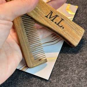 May include: A wooden comb with a folding design. The comb has a brown wood finish and the words "M.L." are engraved on the handle.
