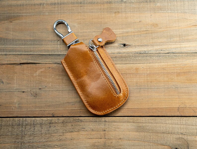 Pu&ograve; includere: Un portachiavi in ​​cuoio marrone con una cerniera argentata e un anello argentato. Il portachiavi &egrave; su una superficie di legno.