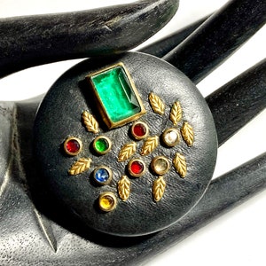 May include: A round, black brooch with a rectangular emerald-colored stone and various colorful gemstones. Gold-colored leaf accents surround the gemstones. The brooch is a unique piece of jewelry.