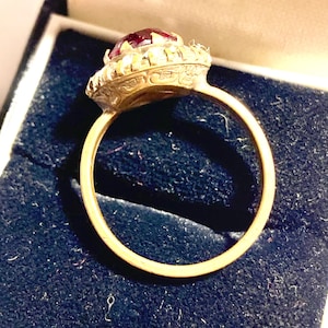 May include: A gold ring with a large, faceted, deep red gemstone at the center. The gemstone is surrounded by smaller, clear stones. The ring is set in a detailed, ornate setting and is displayed in a dark blue jewelry box.