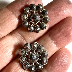 May include: Two silver-toned metal buttons with a raised, faceted design. Each button has a circular shape with a central hole and a decorative border of smaller, raised circles.