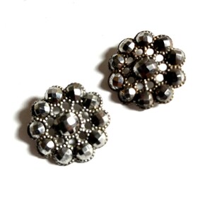 May include: Two silver-toned metal buttons with a flower design. Each button has a circular shape with a raised center and a series of smaller circles around the edge.