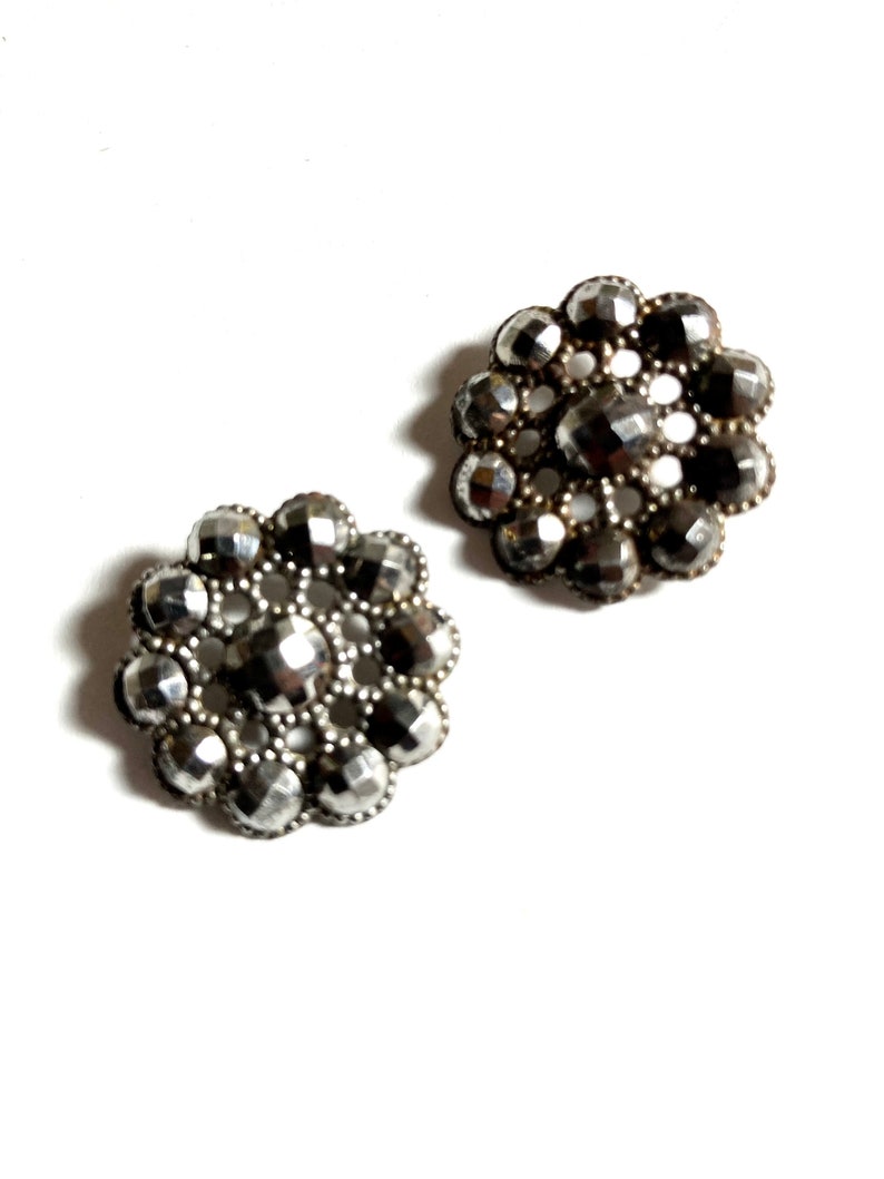 May include: Two silver-toned metal buttons with a flower design. Each button has a circular shape with a raised center and a series of smaller circles around the edge.