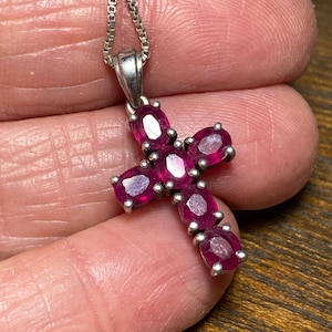 May include: A silver pendant necklace featuring a cross design. The cross is adorned with oval-shaped, deep red gemstones. The necklace chain is a delicate, box-style link.