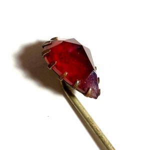 May include: A vintage hat pin featuring a faceted, deep red gemstone. The gemstone is set in a metal frame and mounted on a slender, silver-toned pin. The pin is designed to secure hats or other accessories.