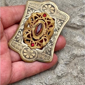 May include: A vintage silver and gold brooch with intricate filigree design and three red glass stones. The brooch has a rectangular shape with rounded corners.