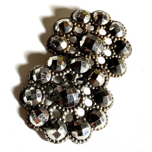 May include: Two silver-toned metal buttons with a flower design. Each button has a circular shape with a raised center and a decorative border of small beads. The buttons are made of a shiny metal with a slightly textured surface.