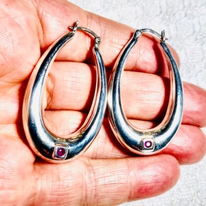 May include: A pair of silver oval hoop earrings with a small pink stone set in each hoop.