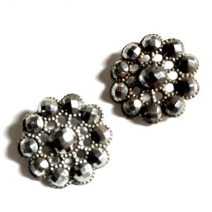 May include: Two silver-toned metal buttons with a flower design. Each button has a center circle surrounded by a ring of smaller circles.