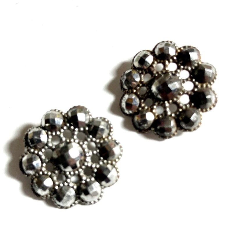 May include: Two silver-toned metal buttons with a flower design. Each button has a center circle surrounded by a ring of smaller circles.