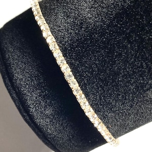 May include: A gold-toned tennis bracelet with a continuous line of clear, sparkling gemstones. The bracelet is displayed on a black velvet surface, creating a contrast that highlights the jewelry's brilliance. The bracelet is a classic piece of jewelry.