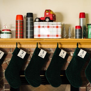 Puede incluir: Seis calcetines de terciopelo verde con etiquetas numeradas colgando de una repisa de chimenea de ladrillo. Los calcetines están decorados con una etiqueta blanca que dice "Santa's Club". La repisa de la chimenea está decorada con un camión de juguete rojo, un termo de cuadros, un termo blanco y negro, una caja de cuadros blancos y rojos y una caja de pan verde.