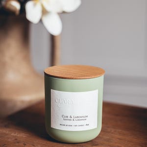 Peut inclure: Une bougie en soja verte avec un couvercle en bois. L'étiquette indique "Clara & Co. Cuir & Labdanum Leather & Labdanum Bougie de Soja - Soy Candle - 8oz"