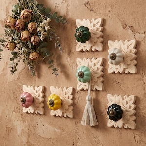 Pode incluir: Uma coleção de ganchos de parede decorativos com costas de madeira entalhada com motivos florais. Cada gancho apresenta um puxador de cerâmica colorido em tons de verde, rosa, amarelo, branco e preto. Um arranjo de flores secas pendura-se perto.