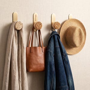 May include: Four wall hooks with a gold finish and woven wood accents. A beige scarf, brown tote bag, denim jacket, and straw hat hang from the hooks. The text "Allhome" is printed on two of the hooks.