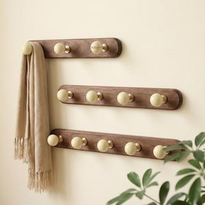 Può includere: Tre attaccapanni in legno con pomelli color crema e ferramenta in ottone sono montati su una parete colorata. Una sciarpa beige è appesa a uno dei pomelli. I supporti sono di diverse lunghezze e sono realizzati in legno scuro. La parola "Allhome" è stampata su uno dei supporti.