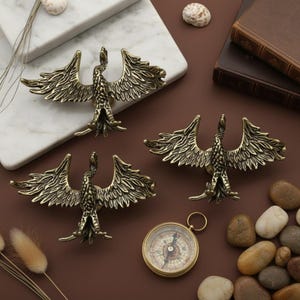 May include: Three bronze-coloured bird-shaped cabinet pulls with outstretched wings. A vintage compass and small stones are also visible. The pulls are on a brown surface with a white marble accent.