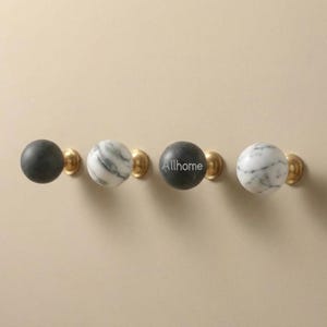 Può includere: Quattro pomelli decorativi per mobili. Due sono in marmo con venature grigie, uno è grigio pieno e uno è grigio pieno con la scritta "Allhome" stampata sopra. Ogni pomello è montato su una base in ottone.