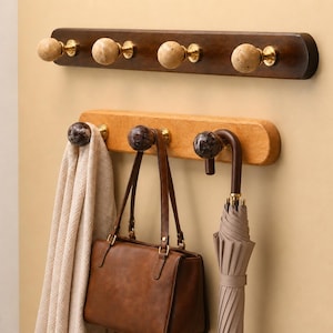 May include: Two wall-mounted coat racks with rounded ends. The top rack is dark brown wood with four beige stone knobs on brass bases. The bottom rack is light brown wood with three dark brown stone knobs, holding a beige scarf, a brown bag, and an umbrella.
