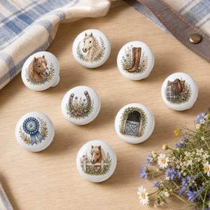 May include: A set of seven round white ceramic cabinet knobs, each featuring a unique equestrian-themed illustration including horses, riding boots, a saddle, a horseshoe, a blue ribbon, a stable gate, and a horse peeking over a wooden fence.
