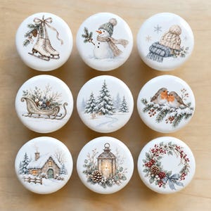May include: A set of nine round white ceramic knobs featuring winter-themed illustrations including ice skates, a snowman, a knit hat and scarf, a sleigh, a snowy forest, two robins, a cottage, a lantern, and a festive wreath with red berries.