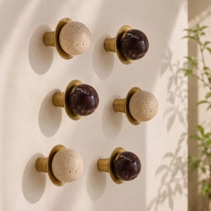 May include: A set of six wall-mounted knobs featuring a brass-toned conical base and a spherical top. Three knobs have a textured, porous beige stone sphere, while the other three feature a smooth, polished dark brown marble sphere.