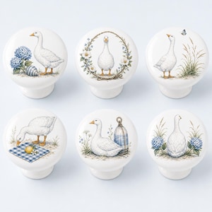 May include: A set of six round white ceramic cabinet knobs, each featuring a unique illustration of a white goose. The designs include geese with blue hydrangeas, floral wreaths, tall grasses, a blue checkered picnic cloth, and nautical buoys.