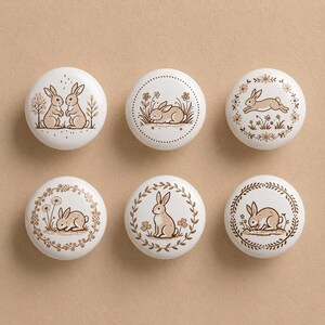 以下が含まれることがあります： 茶色のウサギのイラストが描かれた、白いセラミック製の丸いキャビネットノブが6つ。各ノブには異なるウサギのデザインが施され、花の模様や葉の模様で囲まれています。ノブは丸く、光沢のある仕上がりで、家具に気まぐれな雰囲気を加えるのに最適です。