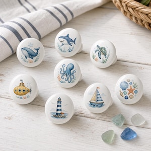 May include: A set of eight round white ceramic cabinet knobs featuring nautical illustrations including a blue whale, a shark, a sea turtle, an octopus, a yellow submarine, a lighthouse, a sailboat, and a collection of seashells and a starfish.