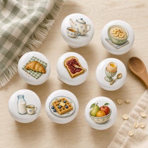 Könnte beinhalten: Ein Set aus acht weißen Keramik-Möbelknöpfen, die jeweils mit einer einzigartigen Frühstücks-Illustration versehen sind, darunter eine Teekanne, Haferbrei, ein Croissant, Toast mit Marmelade, ein weichgekochtes Ei, Milch, Waffeln mit Blaubeeren und eine Schale mit frischem Obst.