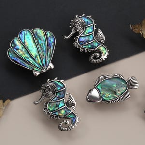 Op de afbeelding: Een verzameling sieraden met oceaanthema, waaronder een schelp, twee zeepaardjes en een vis, allemaal met iriserende abalone schelpinleg en zilverkleurig metaal. Gepresenteerd op een neutrale achtergrond.