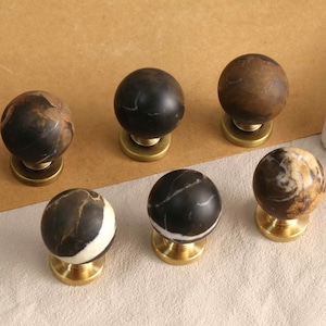 Può includere: Un set di sei pomelli sferici per armadietti con motivi in pietra naturale nelle tonalità del nero, marrone e crema. Ogni pomello è montato su una base metallica circolare color ottone, mostrando venature e texture uniche sulle superfici.