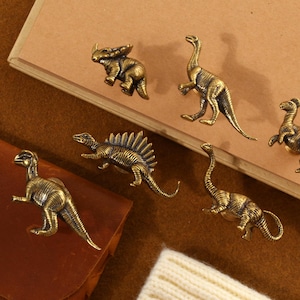 Op de afbeelding: Een verzameling antieke messing dinosaurusbeeldjes, waaronder een triceratops, een brontosaurus en een stegosaurus. De gedetailleerde miniaturen zijn gerangschikt op een bruin leren oppervlak en een notitieboek.
